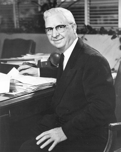 White man with glasses in suit and tie sitting at his desk