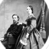 White man with a beard sitting down with young white woman standing up in a dress with curtains behind them