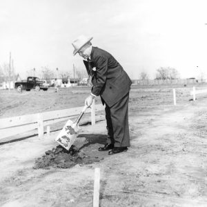 Old white man wearing a hat in a suit using a shovel