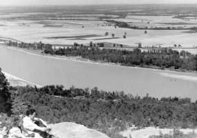 Aerial view of river and fields in the background, white man and woman perched on rock outcropping in foreground