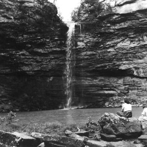 Natural waterfall and pool being observed by two woman sitting on rocks at the edge of the water