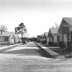 Neighborhood of identical homes with dirt road, sidewalks parked car, and water tower in distance