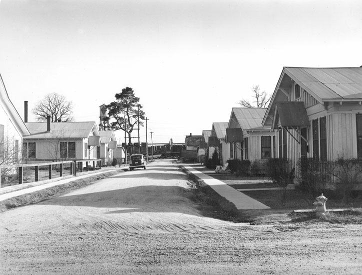 Neighborhood of identical homes with dirt road, sidewalks parked car, and water tower in distance