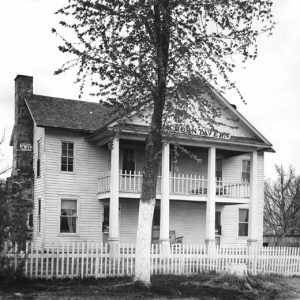 Two-story structure with brick chimney, white picket fence, and tree beyond the fence