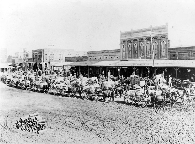 Wagons loaded with cotton crowding dirt road that runs in front of several buildings