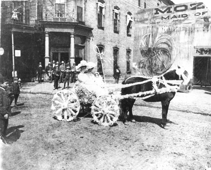 Two white women with little girl in white on a horse drawn buggy with brick building and people in the background