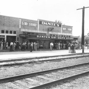 Crowd of people standing outside "Dante's" department store, with railroad tracks in foreground
