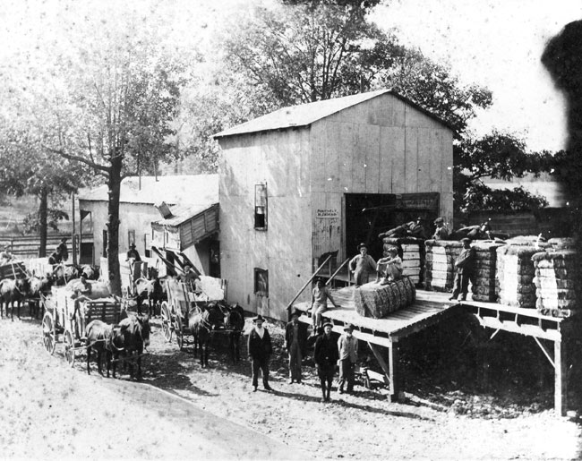Men posing outside barn on loading dock with bundles near horses, wagons
