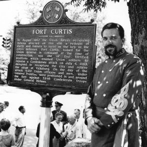 White man in military uniform poses by "Fort Curtis" sign, white people chatting in background