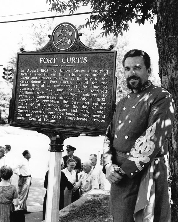 White man in military uniform poses by "Fort Curtis" sign, white people chatting in background