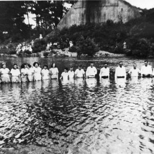 White women, children, and men stand in river with observers, hillside concrete wall in background