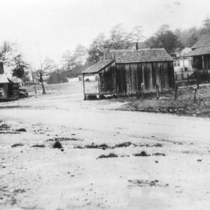 Muddy road, man with mustache, hat on porch, buildings in distance with sign "Dick Huddleston Lum Abner"