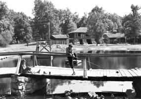 Two white boys fish off wooden bridge with lily pads in foreground, stone lodge in background.