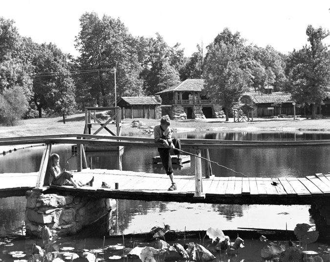 Two white boys fish off wooden bridge with lily pads in foreground, stone lodge in background.
