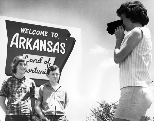 White girl and boy photographed by woman in front of sign "Welcome to Arkansas Land of Opportunity"