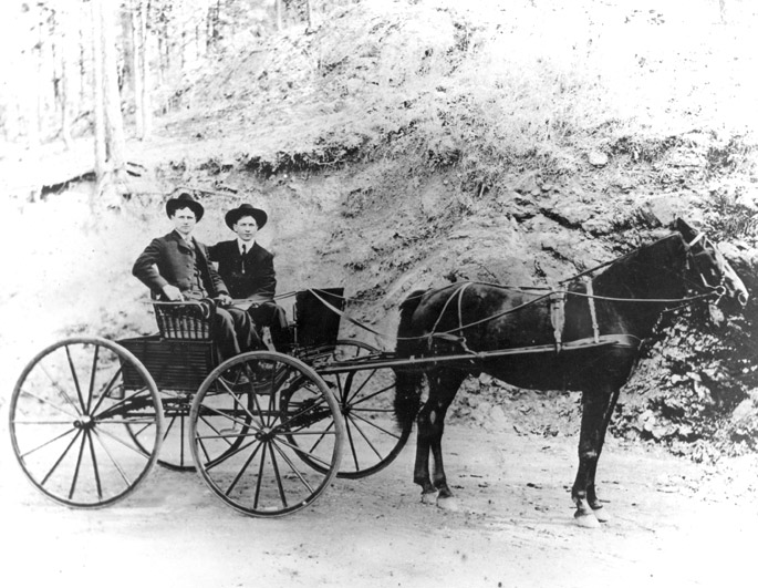Two men pose in suits, hats, on horse-drawn carriage by dirt back slope