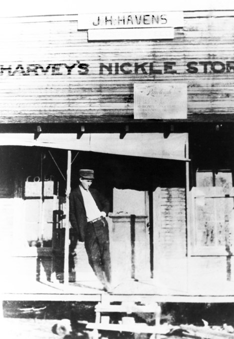 White man in hat hand in pocket leaning on "Harvey's Nickle Store" porch post
