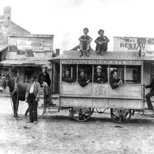 White men pose on horse-drawn trolley labeled "Walnut Ridge & Hoxie, 2" with store, restaurant behind