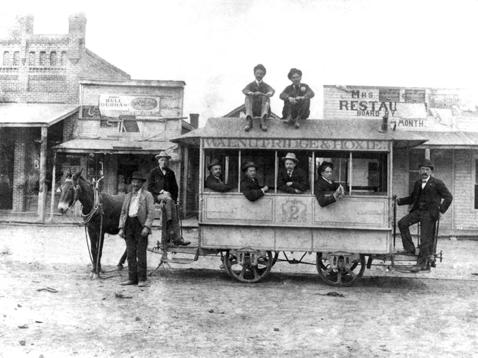 White men pose on horse-drawn trolley labeled "Walnut Ridge & Hoxie, 2" with store, restaurant behind