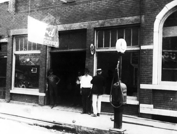 Four men stand by entrance brick gas station with pump and "Ford authorized service" sign