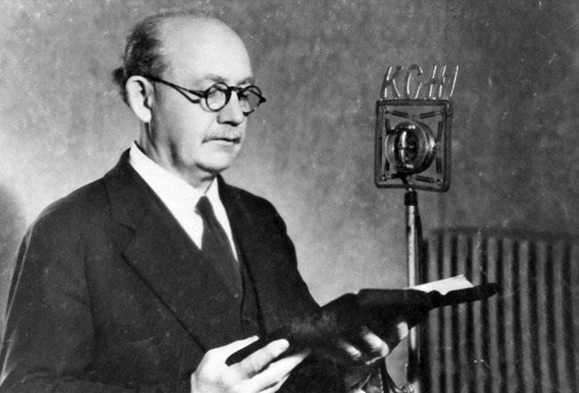 Portrait white man suit, tie, glasses, reading from paper by "KCHI" microphone