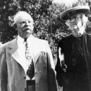 Older white man in suit and woman in hat and dress carrying book, both outdoors