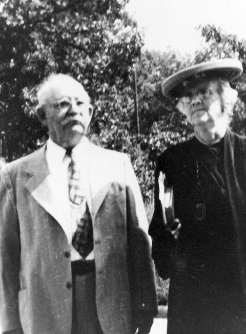 Older white man in suit and woman in hat and dress carrying book, both outdoors