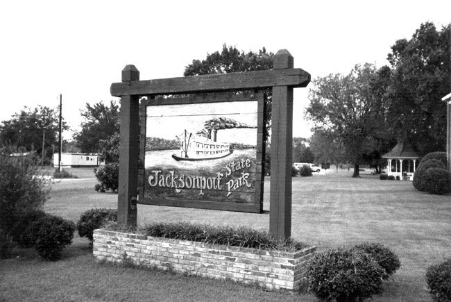 Wooden sign "Jacksonport State Park" with riverboat carving in field, gazebo in background