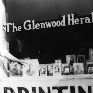 White man in hat, vest, and tie points to soldier portrait in window with signs "The Glenwood Herald, Printing"