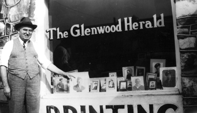 White man in hat, vest, and tie points to soldier portrait in window with signs "The Glenwood Herald, Printing"