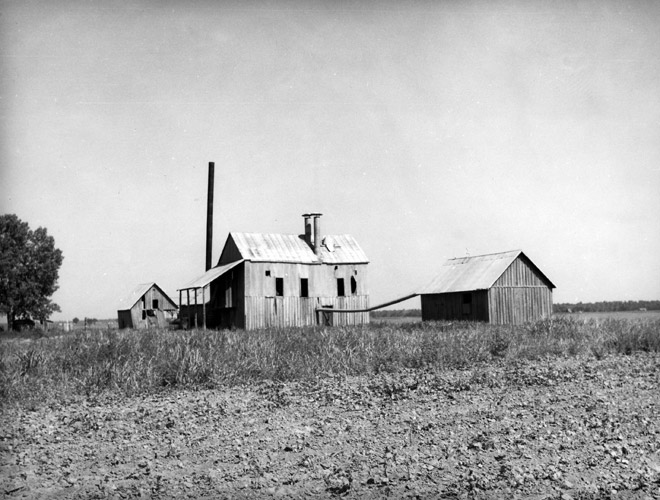 Three barn-like building, wood framed with connecting pipes and chimneys with farm field in foreground