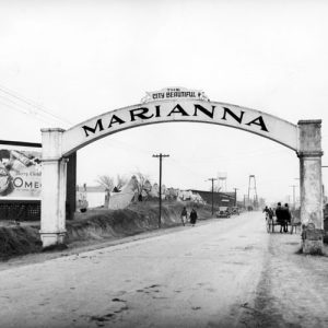 Concrete archway with sign "The City Beautiful Marianna" over road with billboard, pedestrians, tent rows, town in distance