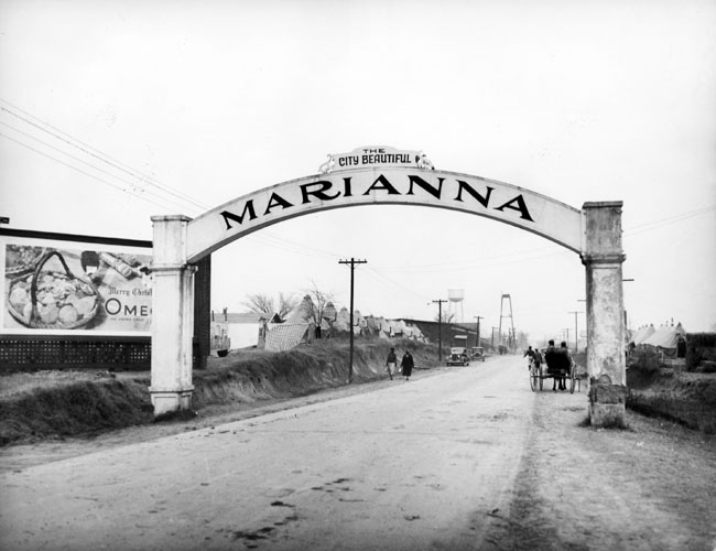 Concrete archway with sign "The City Beautiful Marianna" over road with billboard, pedestrians, tent rows, town in distance