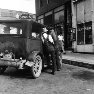 White men in overalls, hats, lean into vehicle by building with signs "Corner Drugs," "Cigars," "Meat Market"