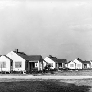 Neighborhood with row of wood framed homes off gravel road, by field
