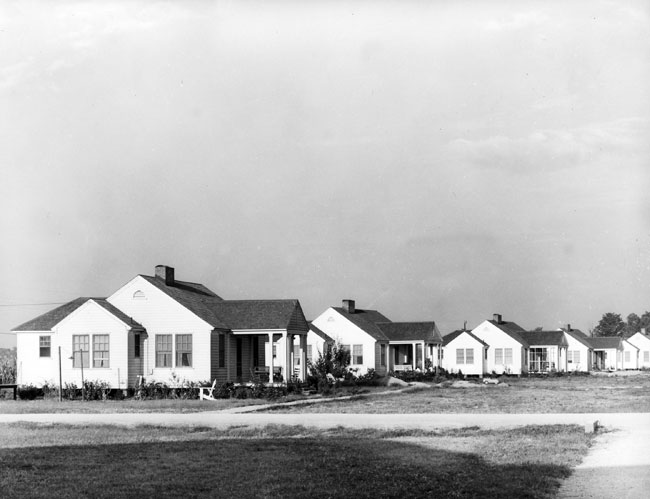 Neighborhood with row of wood framed homes off gravel road, by field
