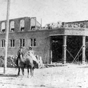 White man and woman pose on horses with brick building under construction in background
