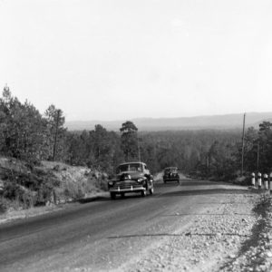Mountain highway with two cars driving uphill past striped posts on gravel shoulder