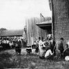 Crowd of people in grass, seated and standing, among brick and wooden buildings
