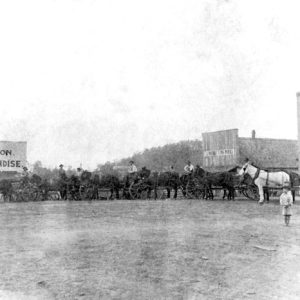 White boy standing near row of men with horses, wagons and banner "I.N. Runyan & Son General Merchandise"