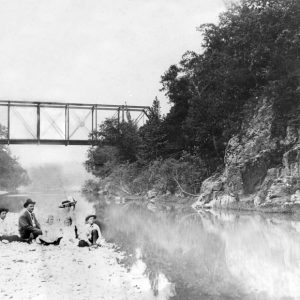 White family sitting on river bank with steel truss bridge behind them