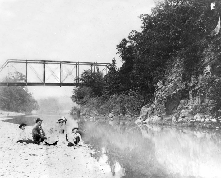 White family sitting on river bank with steel truss bridge behind them