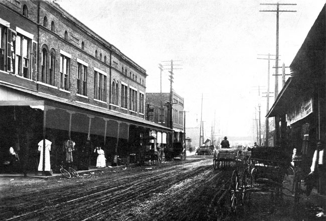 Dirt city street lined with brick architecture, women in dresses below awning, horse drawn carriages, pedestrians