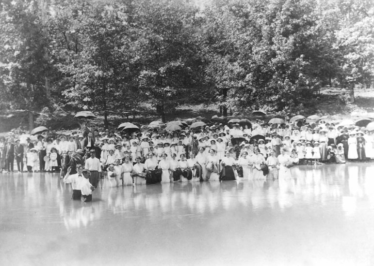 Crowd of people, some with umbrellas, standing by a riverside while others stand in the river