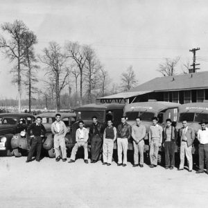 Japanese American men in casual attire pose before cars, ambulances in lot outside wood frame building