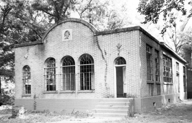 Brick building in woods with large windows arched facade including centered Native American relief portrait