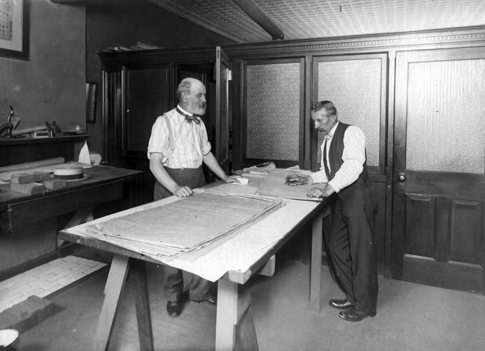 Office two white men examine large sheets paper on sawhorse desk with bricks opposite table