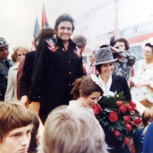 white man in black embroidered alongside white woman in dark suit and hat carrying roses amid a crowd of people