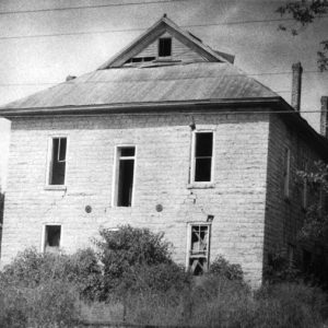 Abandoned two-story building with broken windows and hip roof with overgrowth