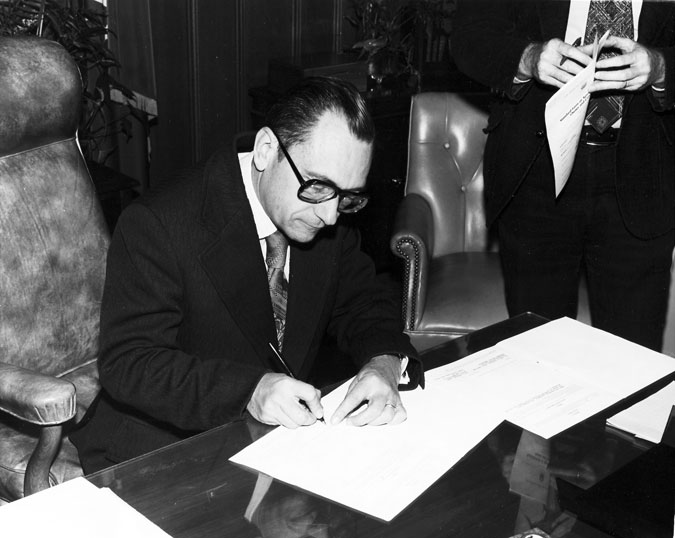 Interior, white man in suit seated signing paper, with white man standing holding form